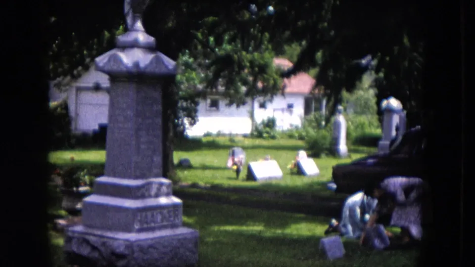 1953 Milwaukee cemetery footage shows two women crouched near a headstone, with a tall monument, lawn, trees, and nearby grave markers visible.
