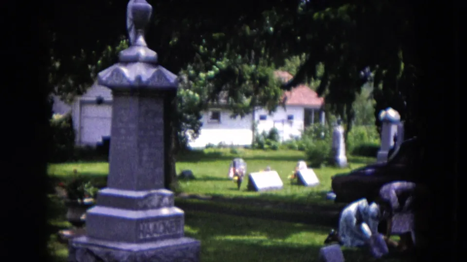 1953 Milwaukee 8mm still with a tall gray cemetery monument in the foreground and two women tending a gravesite at the right edge.