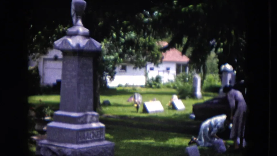 1953 Milwaukee frame showing one woman standing and another bending near a grave marker, with a gray monument and sunlit cemetery lawn nearby.