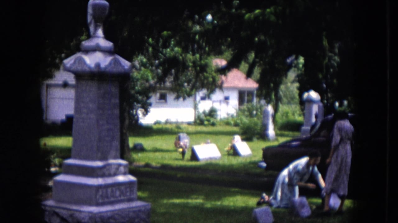 1953 Milwaukee home-movie frame showing two women in a cemetery, with one kneeling near a grave marker and monuments around them.