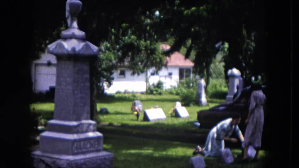 1953 Milwaukee home-movie frame showing two women in a cemetery, with one kneeling near a grave marker and monuments around them.