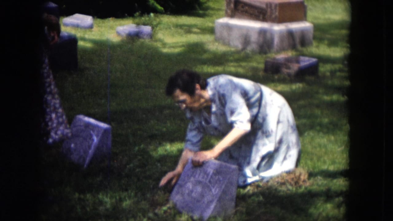 Woman kneeling in grass beside a tilted gravestone in a cemetery.
