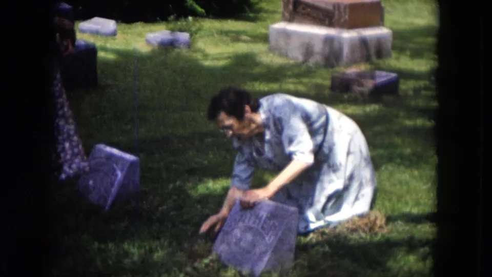 In 1953 Milwaukee home-movie footage, a woman kneels beside a tilted gravestone with other cemetery markers behind her.