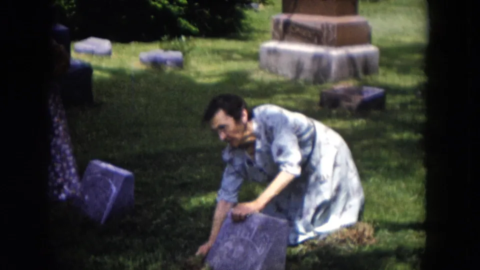 A woman in a light patterned dress reaches toward a gravestone in a Milwaukee cemetery, with monuments visible in the background.