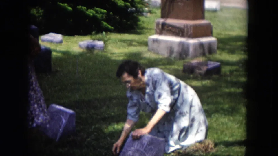 The home-movie frame shows the woman bending over a gravestone, surrounded by grass, shade, and several cemetery markers.