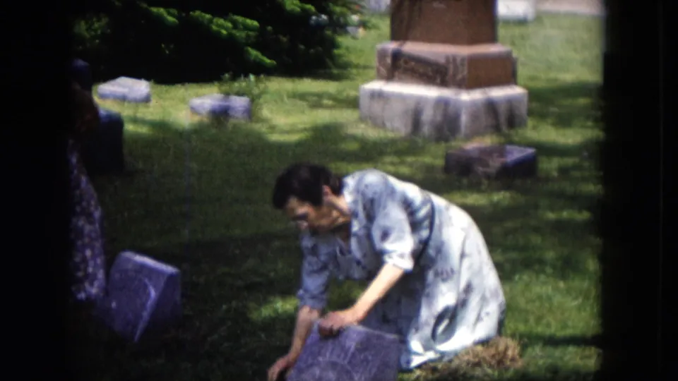 A woman kneels low by a gravestone in the cemetery, with dappled sunlight, headstones, and a larger monument behind her.