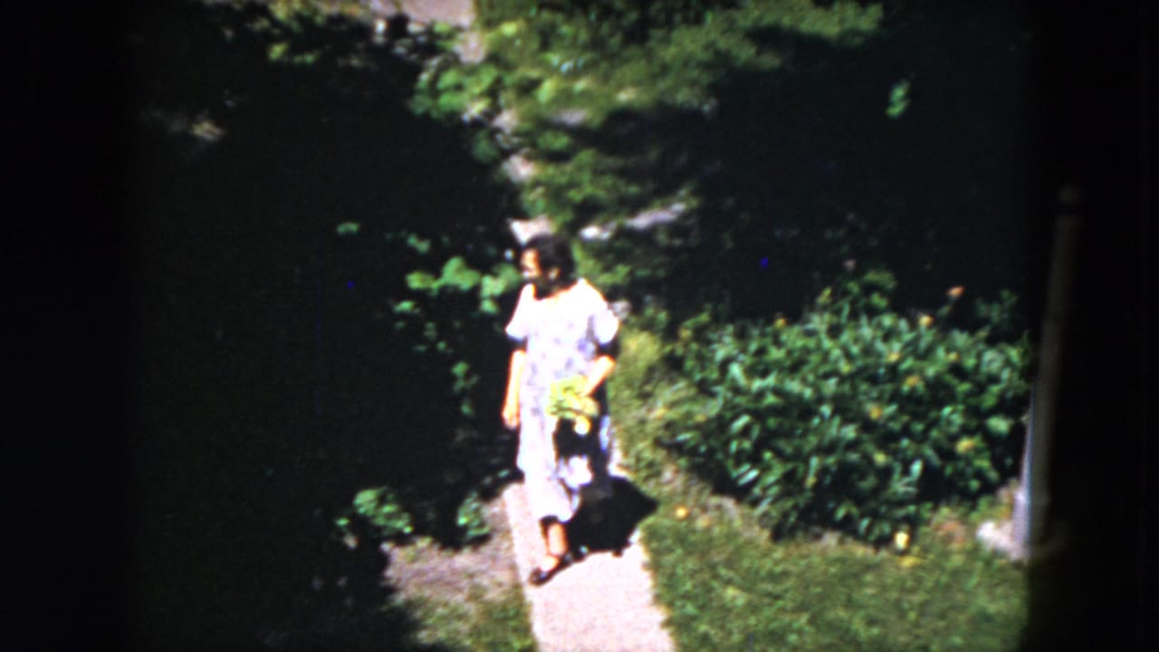 Woman in a light dress walks on a narrow garden path carrying a yellow basket.