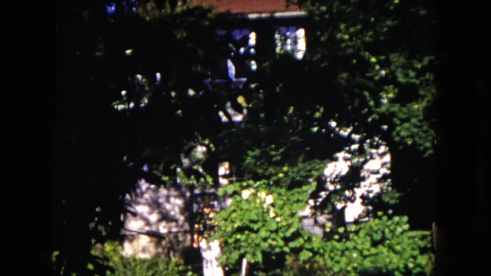 1953 Milwaukee home-movie frame showing a woman in a light dress walking through dense garden foliage near a house.