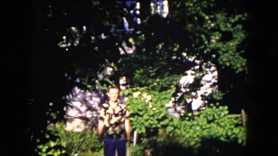 The adult man is closer in frame, wearing a patterned short-sleeve shirt as he walks through the sunlit and shaded garden.