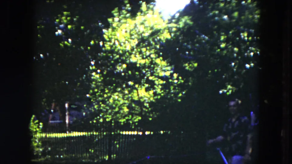 1953 Milwaukee 8mm frame of a sunlit garden and fence, with a person in patterned clothing partly visible at right.