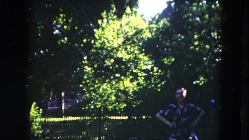 A man in a dark short-sleeve shirt stands with hands on hips near a fence in 1953 Milwaukee park footage.