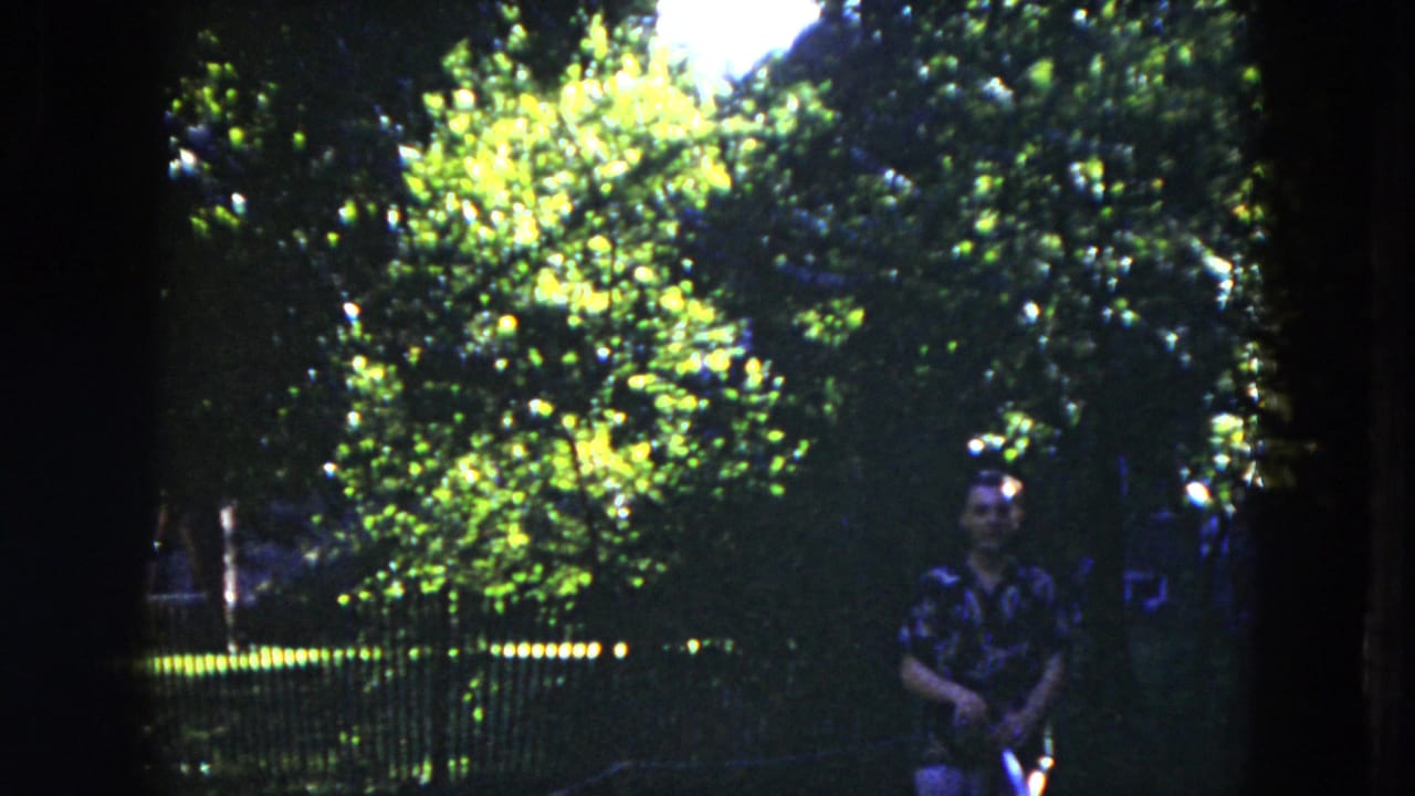 In 1953 Milwaukee home-movie footage, a man in a patterned shirt stands among trees near a dark fence.