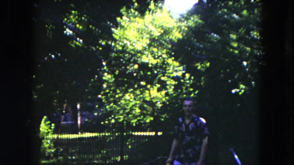 A man appears at the edge of a leafy park or yard in Milwaukee, with bright foliage and a fence behind him.