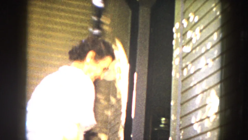 A woman in a light dress leans at a doorway beside a slatted exterior wall with sun patterns across the surface.