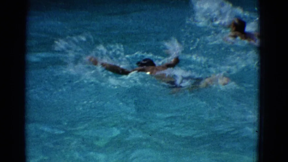 1953 Yonkers home-movie frame showing a swimmer in a dark cap moving through a pool with splashes around the arms.