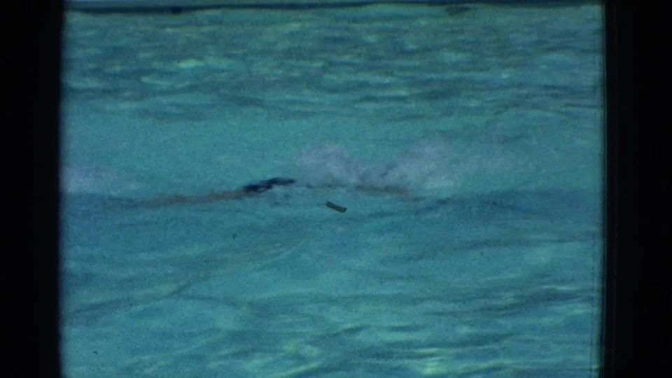 A swimmer in dark shorts moves across the blue pool, with spray and ripples visible in the 1953 home-movie frame.