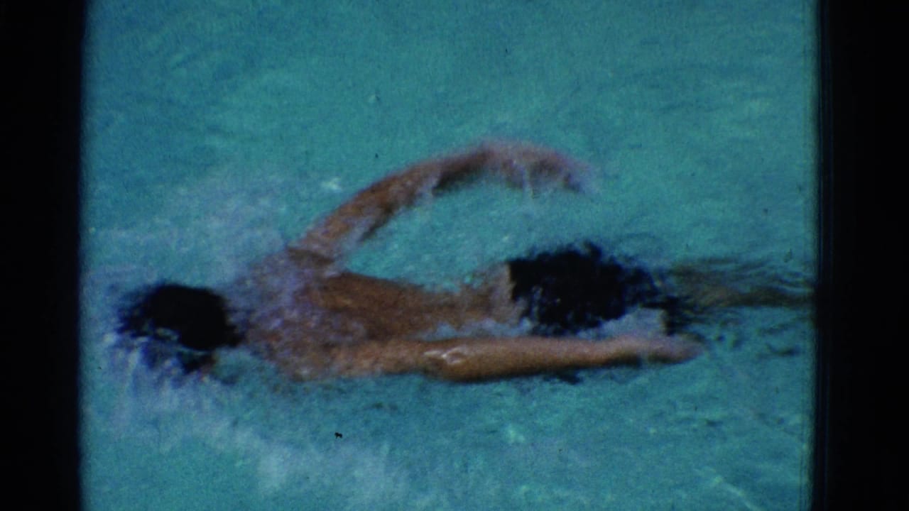 Swimmer in blue trunks moving through teal pool water with one arm raised.