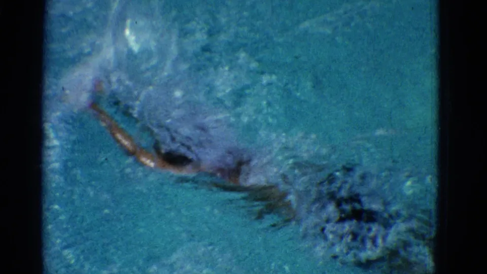 1953 Yonkers pool footage shows the swimmer partly underwater during a forceful stroke, surrounded by splashing water.