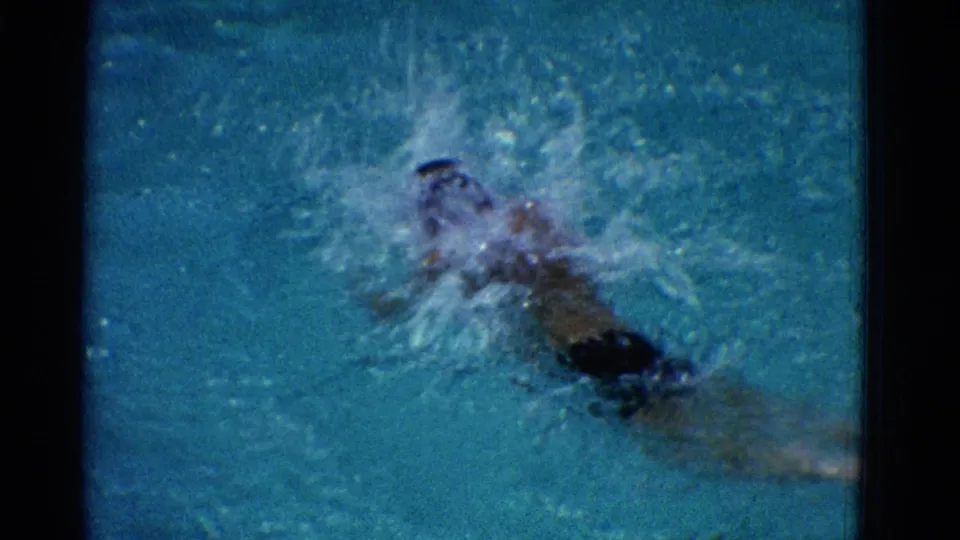 1953 Yonkers home-movie frame with a swimmer advancing through the pool as spray breaks across the face and shoulders.