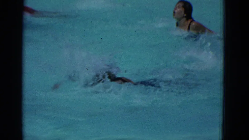 A Yonkers pool race moment shows one swimmer at the surface and another partly obscured by splashing water in grainy 8mm footage.