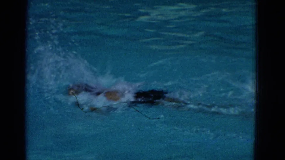 The 1953 home-movie frame shows a swimmer in dark swimwear driving through blue pool water, partly blurred by motion and spray.