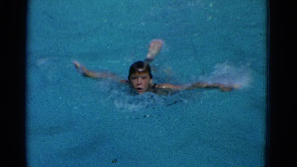 A young swimmer faces the camera with arms spread in blue pool water in supported 1953 Yonkers 8mm home-movie footage.