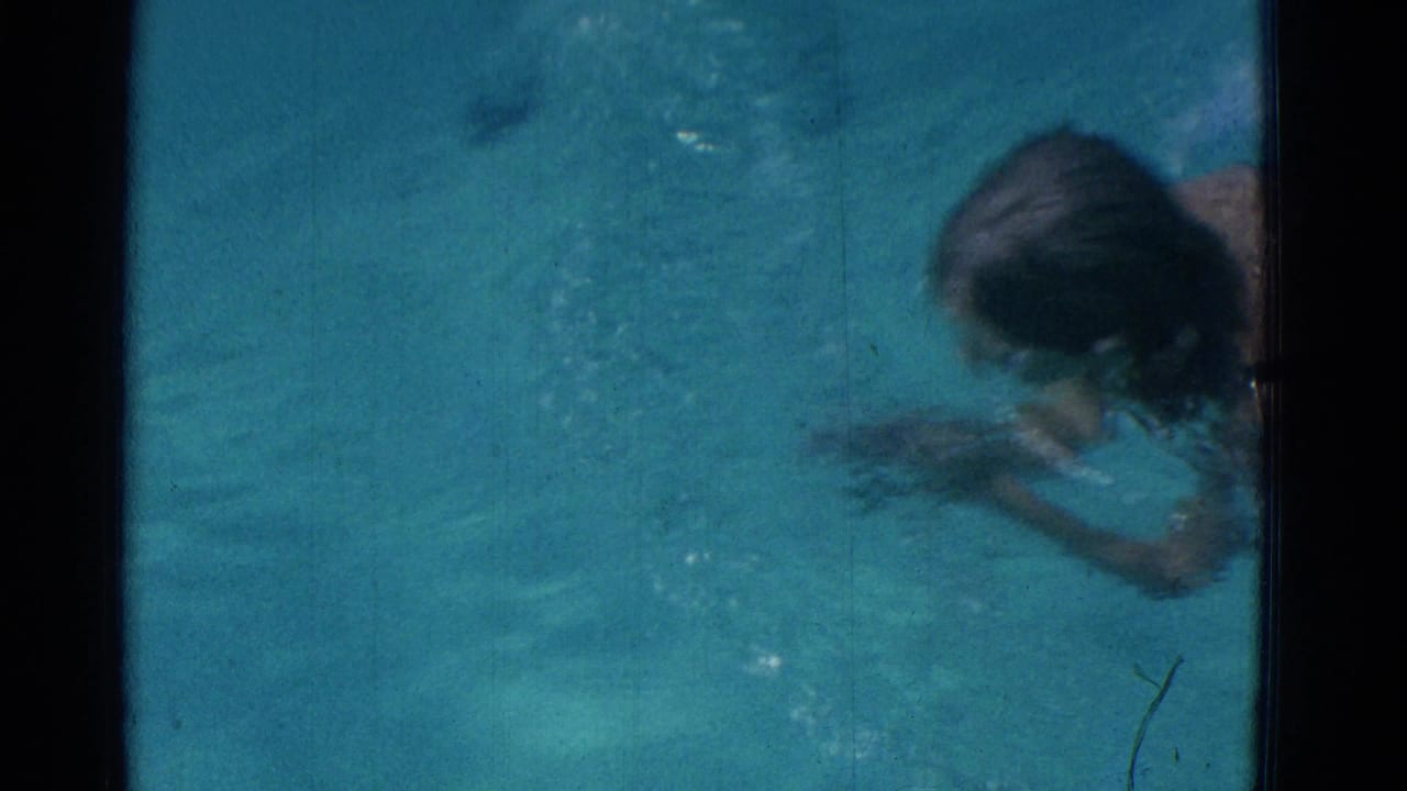 In 1953 Yonkers home-movie footage, a young swimmer in a dark suit moves across blue pool water during a breaststroke motion.