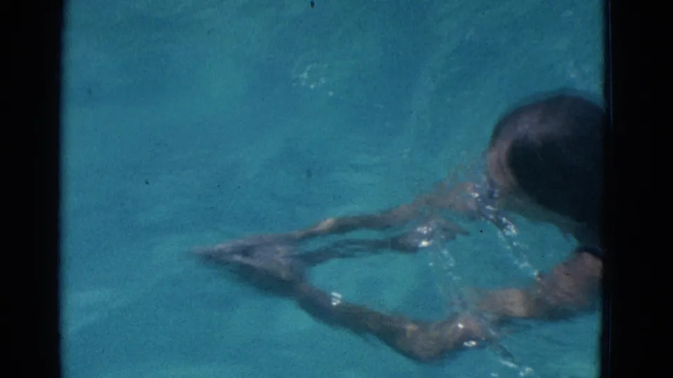 The swimmer continues across the blue pool, with arms forward, legs stretched behind, and surface ripples around her body.