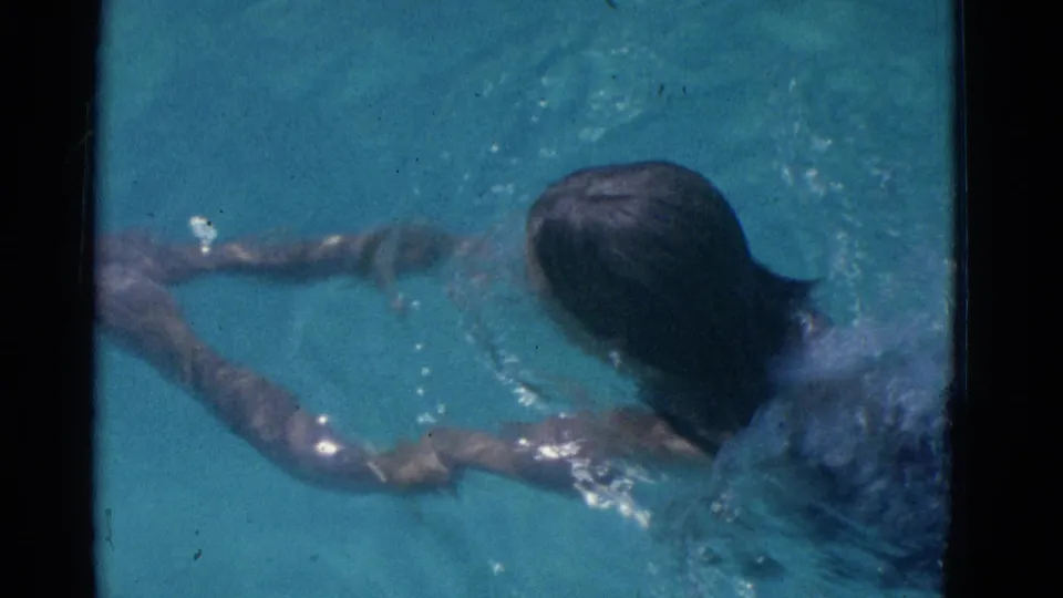 From an overhead home-movie angle, the swimmer's head, shoulders, and forward-reaching arms are visible in the blue pool water.