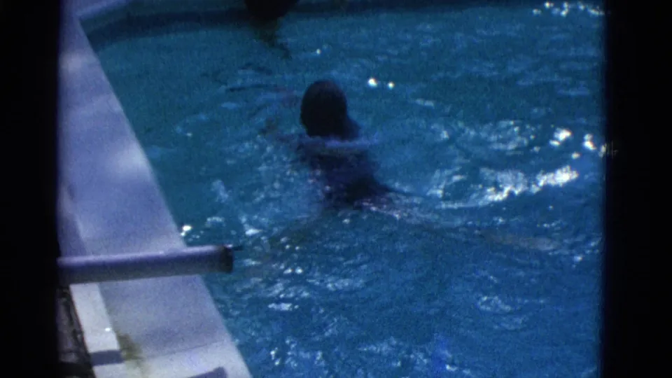 1953 Yonkers pool frame showing a swimmer from behind moving across blue water near a pale pool edge and metal railing.
