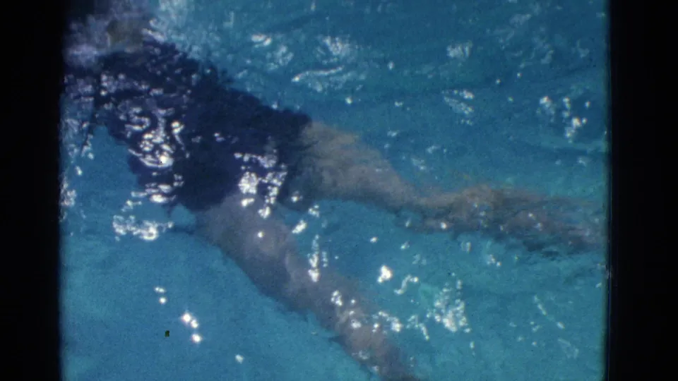 The swimmer paddles across the pool, partly submerged, with bright surface reflections in the 8mm frame.