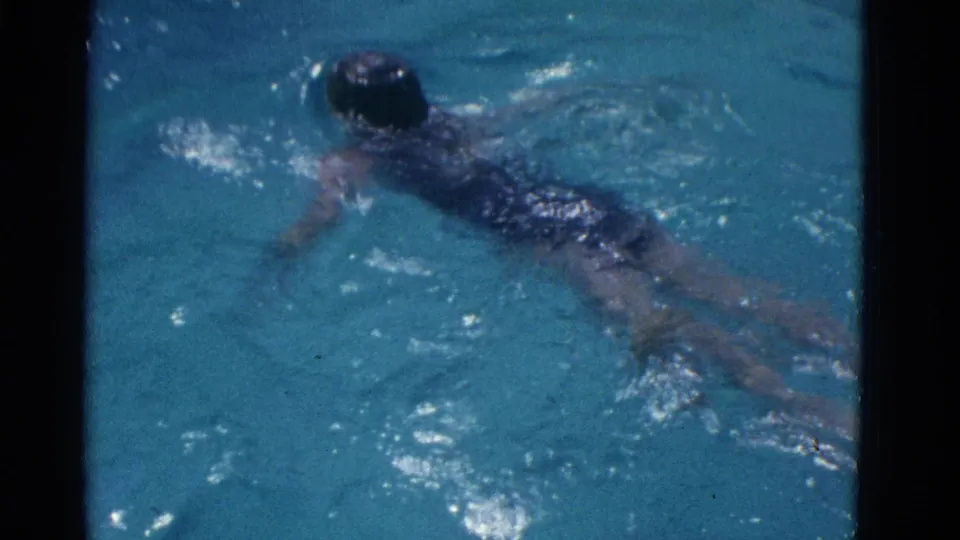 In 1953 Yonkers home-movie footage, a girl in a dark swimsuit swims near the surface of clear blue pool water.