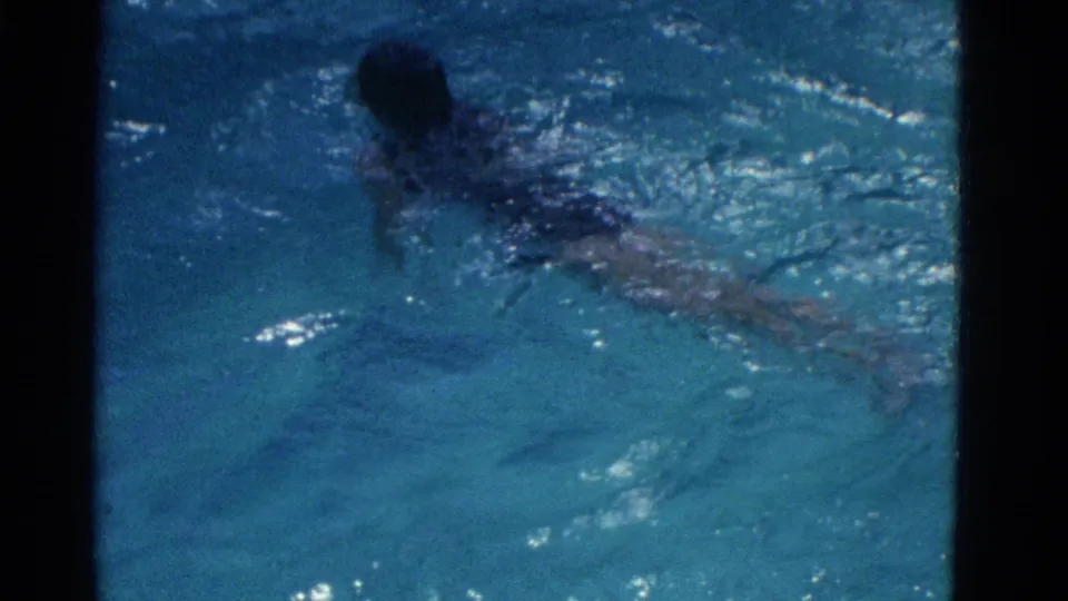 A girl swims through blue pool water from an overhead 8mm home-movie angle, with her body partly softened by the surface.