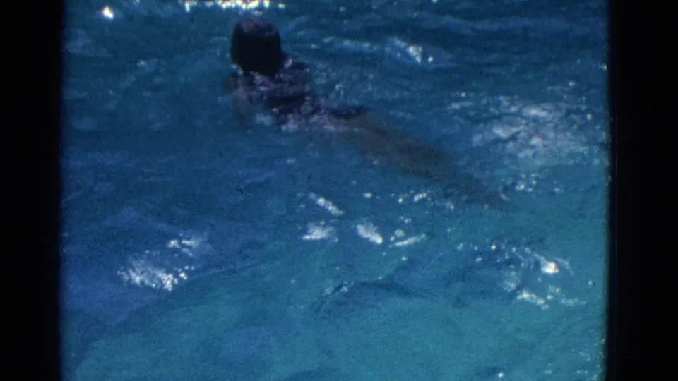 The girl swims away from the camera in blue pool water, her head and shoulders visible through ripples in the 1953 home movie.