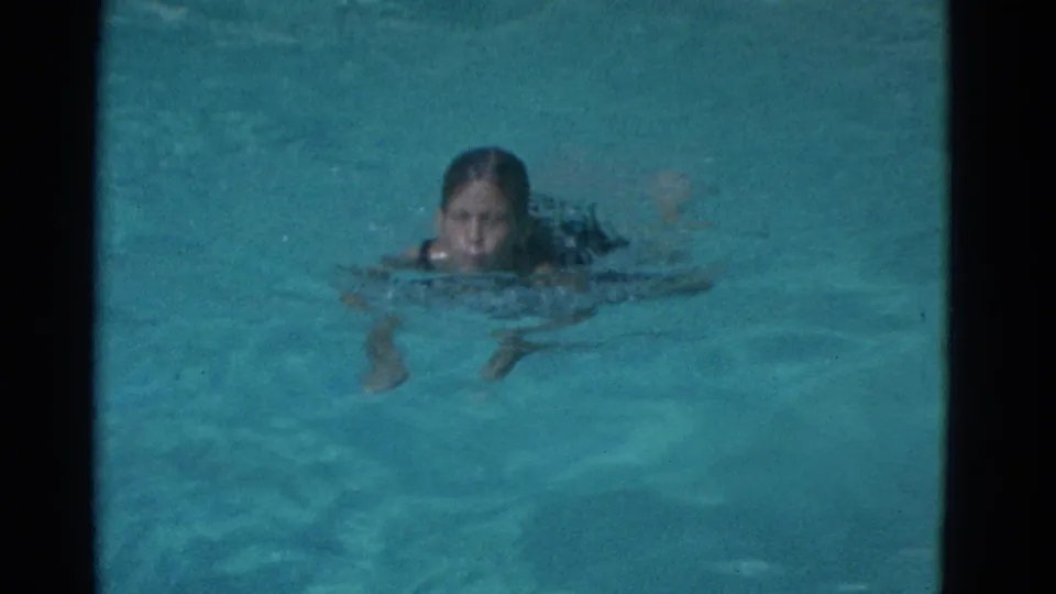 A girl faces the camera while swimming in pale blue pool water, with her face visible above the surface in the Yonkers home movie.