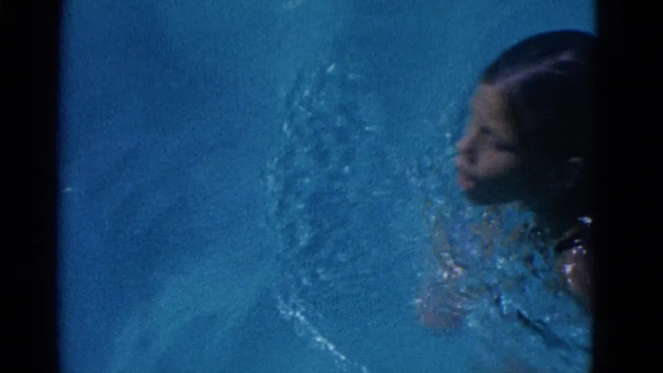 In 1953 Yonkers home-movie footage, a swimmer moves through blue pool water with face, hair, shoulders, and splashes visible.