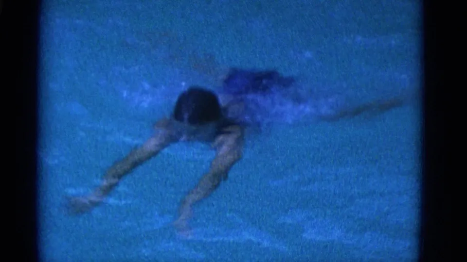 1953 Yonkers 8mm frame of a swimmer in a dark swimsuit moving through blue pool water with arms stretched ahead.