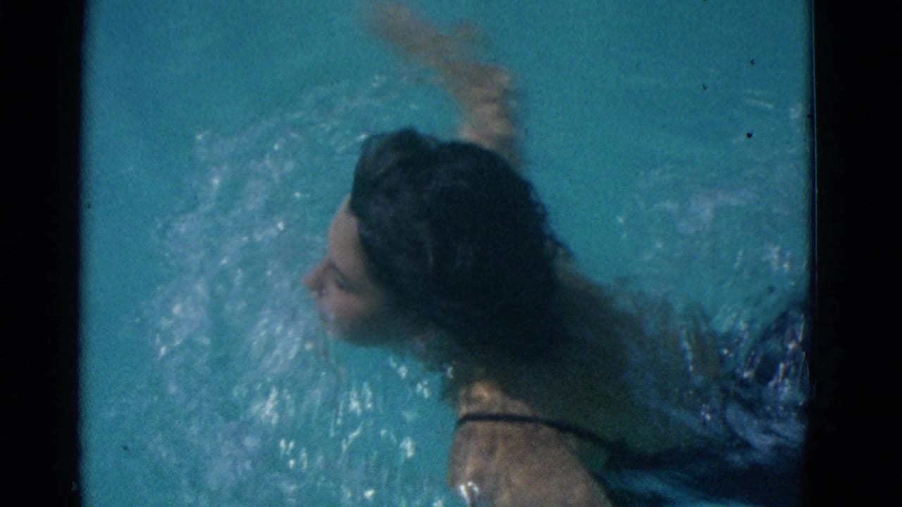 1953 Yonkers home-movie frame showing a swimmer in a dark bathing suit gliding through clear blue pool water from an overhead angle.