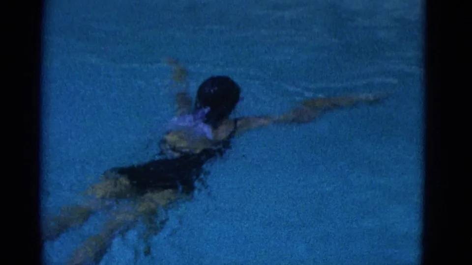 1953 Yonkers home-movie frame showing a swimmer in a dark swimsuit gliding through blue pool water with one arm extended.