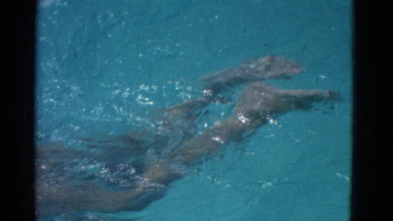 In 1953 Yonkers home-movie footage, a woman in a swimsuit moves through blue pool water, partly softened by grain and exposure.