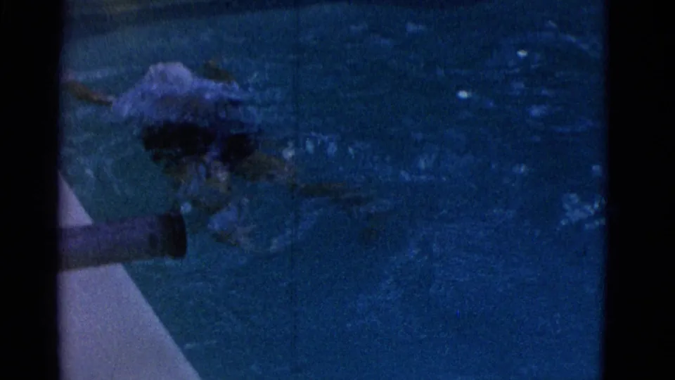 The woman swims near a pool edge in the Yonkers 1953 home movie, with her head and upper body partly obscured by dark blue water and splash.