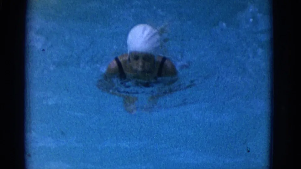 A woman wearing a white swim cap and dark swimsuit faces the camera while treading water in the 1953 Yonkers pool footage.