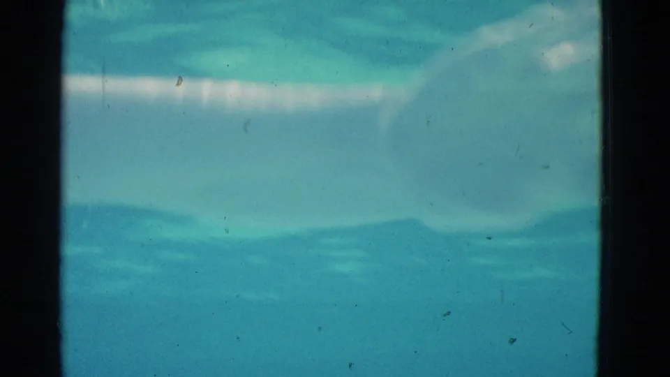 Close Yonkers home-movie frame showing turquoise water, a faint submerged form, surface ripples, dust, scratches, and dark 8mm borders.