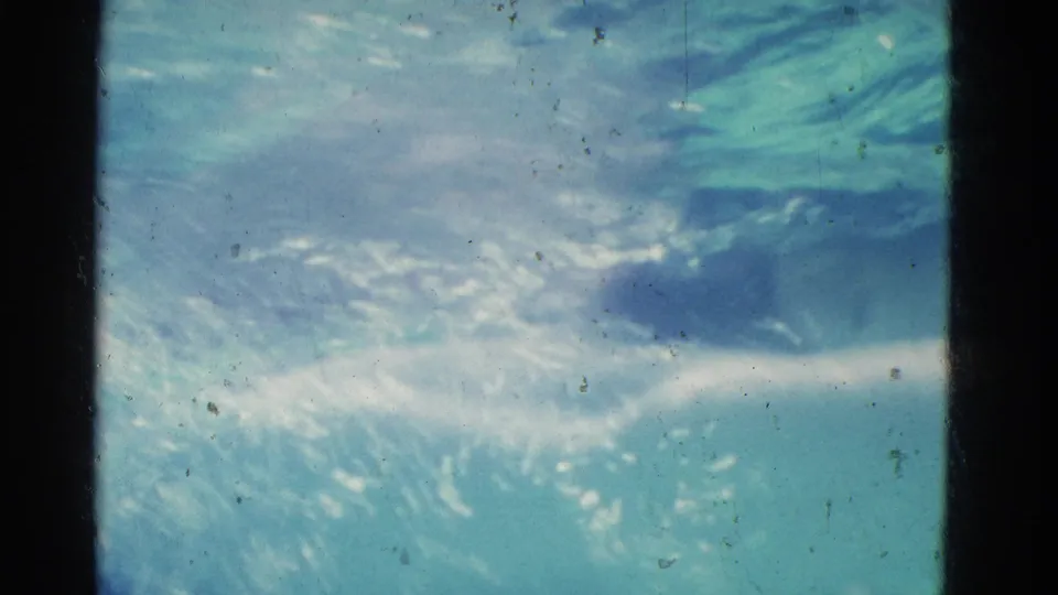 Underwater 8mm frame from the Yonkers clip with rippled sunlight, turquoise water, and faint swimmer movement near the surface.