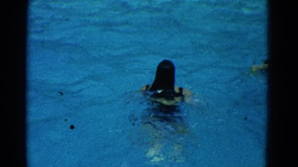 A woman swims away from the camera at the pool surface, with ripples and dark hair visible in the blue water.