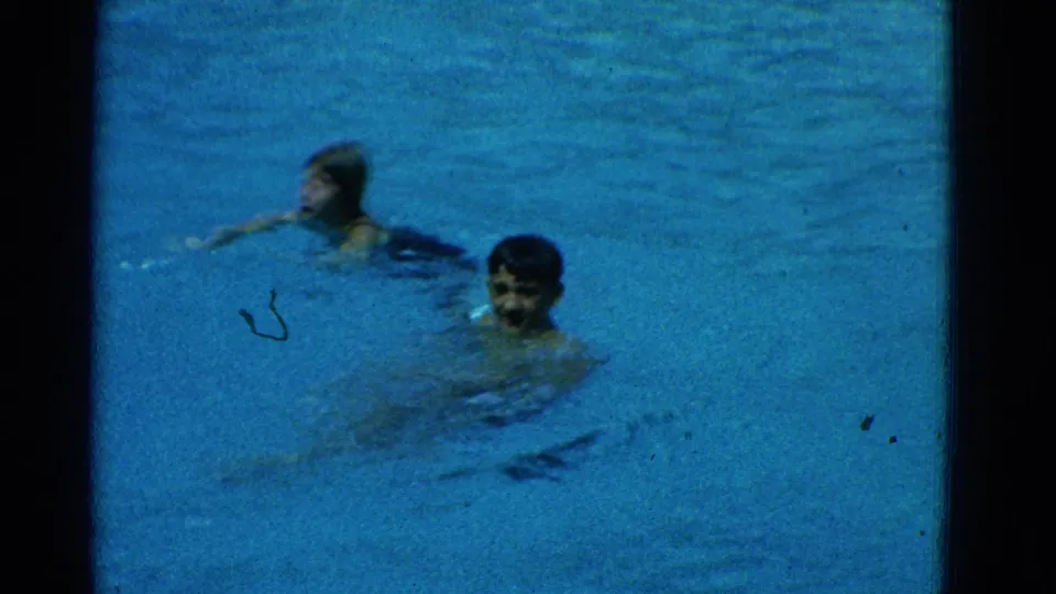 1953 Yonkers home-movie frame with two swimmers paused in the pool, facing toward the camera in blue water.
