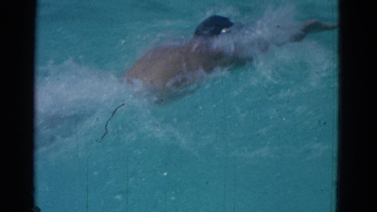 1953 Yonkers home-movie frame showing a swimmer in a dark cap mid-stroke, partly obscured by splashing pool water.