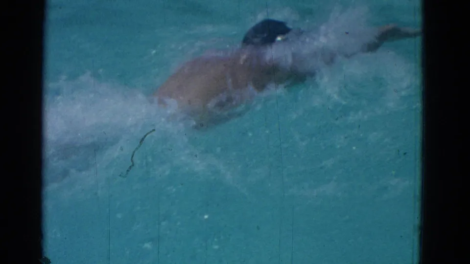 1953 Yonkers home-movie frame showing a swimmer in a dark cap mid-stroke, partly obscured by splashing pool water.