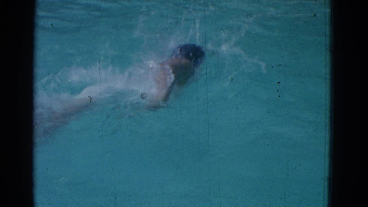 Swimmer in a dark cap moves through turquoise pool water with splashes around the arms.