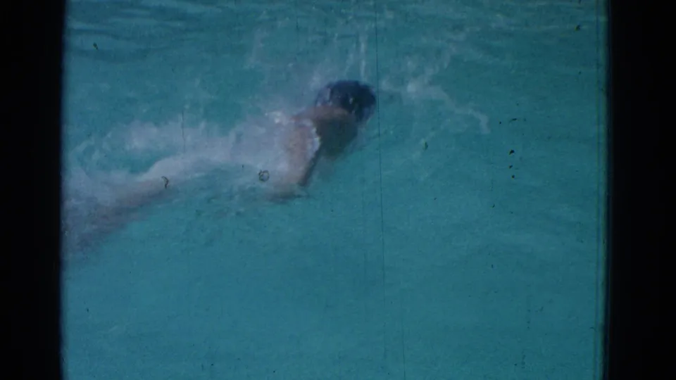 1953 Yonkers 8mm frame showing a swimmer in a dark cap doing freestyle through turquoise pool water with visible film grain and scratches.